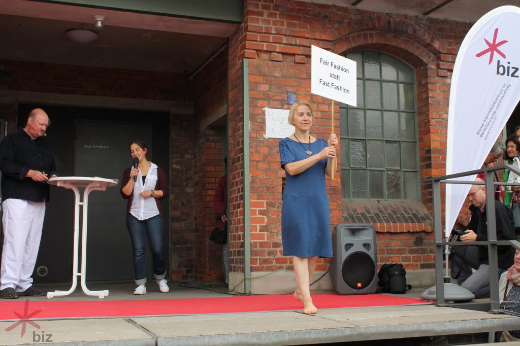Frau mit Protestschild mit zwei Moderatoren im Hintergrund während der Öko-fairen Modenschau