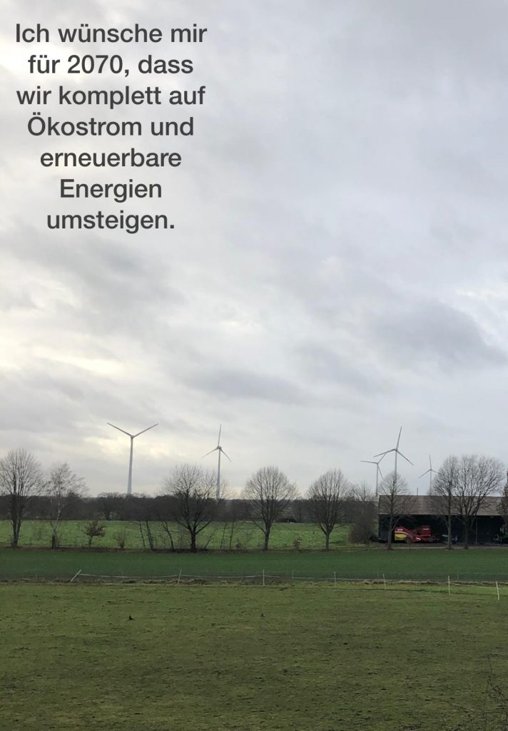 Norddeutsche Landschaft mit Windkraftanlagen im Hintergrund und ein Text mit geschriebenen Wunsch nach einem Umstieg auf Ökostrom bis 2070