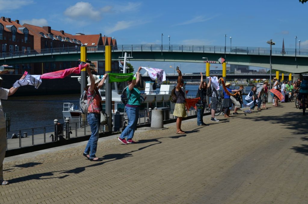 Menschen machen mit Plakaten eine La-Ola-Welle am Rande des Weserweges an der Schlachte