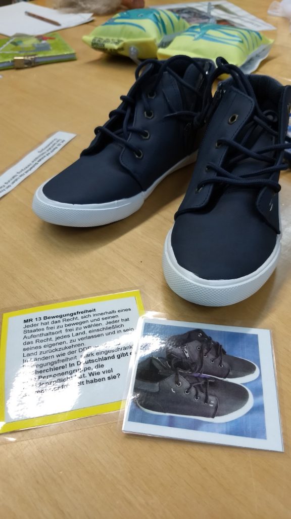 Schuhe und ein Erklärungsschild aus dem Menschenrechtskoffer stehen als Lehrmaterial auf einem Tisch